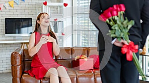 Asian young beautiful female surprised girlfriend in red dress sitting smiling on cozy sofa while unrecognizable male boyfriend