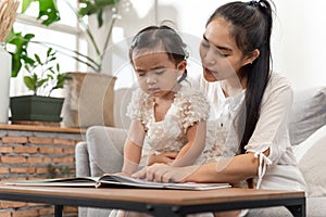 Asian young beautiful mother and daughter sitting on sofa and reading a book story with happiness