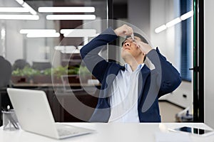 Asian young businessman instills his eyes with medicine drops for inflammation and overfatigue. He sits at his desk in