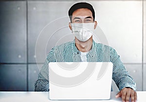 Asian Young Businessman Wearing Surgical Mask in Office. Sitting on Desk while Working on Computer Laptop. Smiling and Looking at