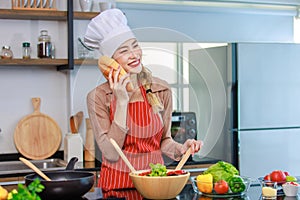 Asian young cheerful female housewife chef wearing white tall cook hat and apron standing smiling holding baguette bread and