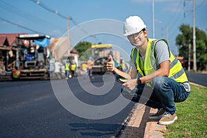 Asian young civil engineer showing confidence with thumb raised at road construction site with Asphalt paver & road roller & dump