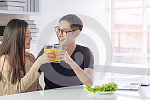 Asian young couple holding a glass of fresh orange juice for health with vegetable salad, laptop, and a smartphone