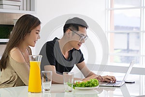 Asian young couple looking at a laptop and smiling while sitting on chair having breakfast in a kitchen at home. Loving couple a