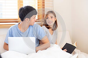 Asian young couple with man using laptop computer and searching internet and woman reading notebook on bed