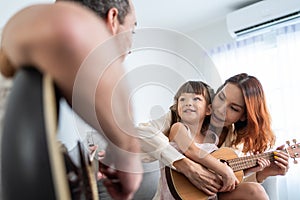 Asian young couple playing guitar with young baby kid together at home. Attractive beautiful parents sit on sofa, spend leisure