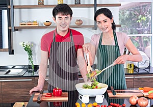 Asian young couple`s in apron, make cooking together. Woman mix salad dressing with vegetable in big bowl.