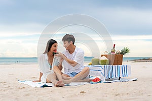 Asian young couple sitting on the picnic blanket and eating watermelon in the beach and near sea with tropical fruit in background