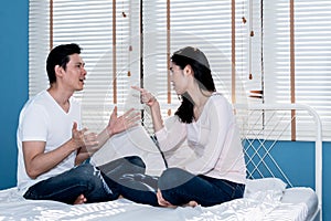 Asian young couple are sitting on the white bed, The attractive wife is tightening her husband to tell the truth