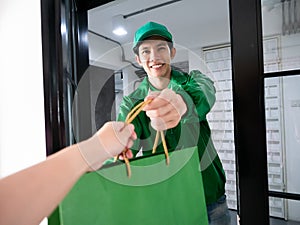 Asian young delivery man in green uniform jacket smile and send shopping paper bag to customer who purchased order online look