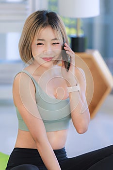 Asian young female athlete teenager in sportswear sport bra sitting smiling on yoga pilates mat holding using smartphone in hand