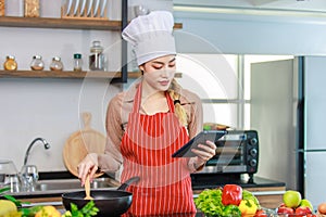 Asian young female chef housewife wears white tall cook hat and apron learning cooking homemade food meal online via touchscreen