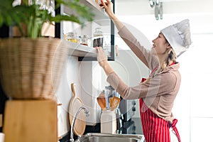 Asian young female chef housewife wears white tall cook hat and apron smiling standing reaching picking up ingredients and dishes