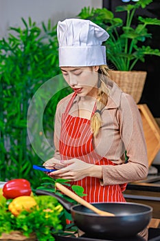 Asian young female chef housewife wears white tall cook hat and apron standing smiling on smartphone call holding wooden spoon