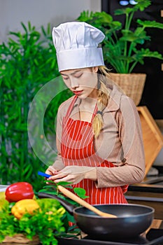 Asian young female chef housewife wears white tall cook hat and apron standing smiling on smartphone call holding wooden spoon