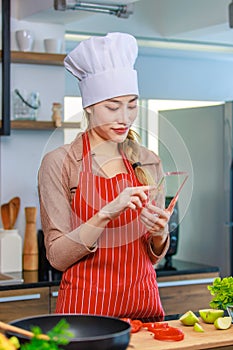 Asian young female chef housewife wears white tall cook hat and apron standing smiling on smartphone call holding wooden spoon