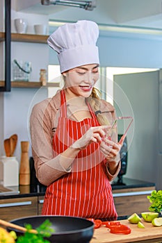 Asian young female chef housewife wears white tall cook hat and apron standing smiling on smartphone call holding wooden spoon