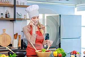 Asian young female chef housewife wears white tall cook hat and apron standing smiling on smartphone call holding wooden spoon