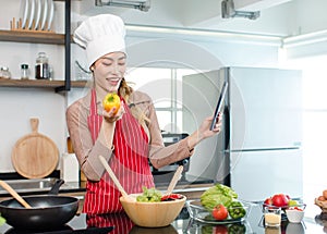 Asian young female chef housewife wears white tall cook hat and apron standing smiling on smartphone call online mixing delicious