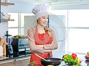 Asian young female chef wears white tall cook hat and apron smiling posing ready to cooking food with pan at counter with bread