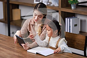 Asian young female housewife mother tutor teacher sitting smiling on table in living room holding tablet computer pointing