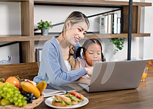 Asian young female mother and little cute girl daughter sitting smiling learning studying doing homework online via laptop