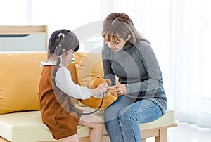 Asian young female mother sitting on cozy sofa smiling holding teddy bear doll playing doctor patient with little cute daughter gi