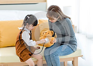 Asian young female mother sitting on cozy sofa smiling holding teddy bear doll playing doctor patient with little cute daughter gi