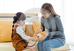 Asian young female mother sitting on cozy sofa smiling holding teddy bear doll playing doctor patient with little cute daughter gi