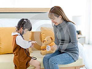 Asian young female mother sitting on cozy sofa smiling holding teddy bear doll playing doctor patient with little cute daughter gi