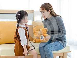 Asian young female mother sitting on cozy sofa smiling holding teddy bear doll playing doctor patient with little cute daughter gi