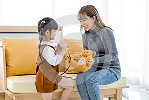 Asian young female mother sitting on cozy sofa smiling holding teddy bear doll playing doctor patient with little cute daughter gi