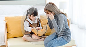 Asian young female mother sitting on cozy sofa smiling holding teddy bear doll playing doctor patient with little cute daughter gi