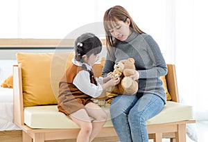 Asian young female mother sitting on cozy sofa smiling holding teddy bear doll playing doctor patient with little cute daughter gi