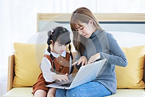 Asian young female teenager mother nanny babysitter sitting on cozy sofa couch helping teaching homework via laptop computer while