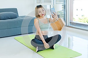 Asian young fit female athlete teenager in sportswear sport bra sitting smiling on yoga pilates mat holding smartphone in hand