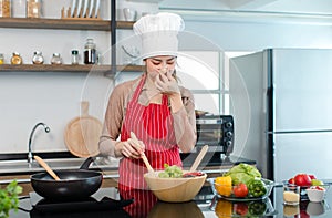 Asian young frowning face female housewife chef wearing white tall cook hat and apron standing holding hand closing nose mixing