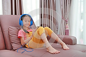 Asian young girl child wearing headphone and using smartphone on sofa in living room at home