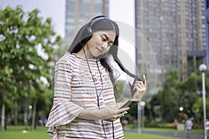 Asian young girl stands listening to music from a smartphone using headphones and copy space. At public park the city center happy