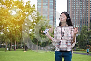 Asian young girl stands listening to music from a smartphone using headphones. At public park the city center happy and relax On