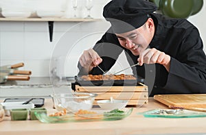 Asian young handsome chef man, in black uniform, smiling while cooking japanese food called takoyaki in hot pan with ingredients