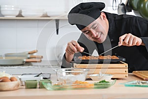 Asian young handsome chef man, in black uniform, smiling while cooking japanese food called takoyaki in hot pan with ingredients