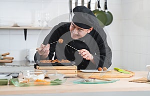 Asian young handsome chef man, in black uniform, smiling while cooking japanese food called takoyaki in hot pan with ingredients