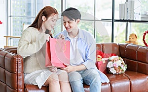 Asian young handsome male boyfriend sitting smiling with beautiful female girlfriend giving red wrapped present gift box in paper