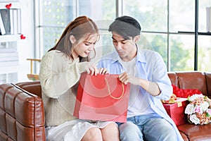 Asian young handsome male boyfriend sitting smiling with beautiful female girlfriend giving red wrapped present gift box in paper