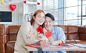 Asian young handsome male boyfriend sitting on sofa smiling cuddling with beautiful female girlfriend holding red heart shape