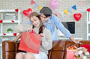 Asian young handsome male boyfriend standing smiling behind beautiful female girlfriend giving red wrapped present gift box in