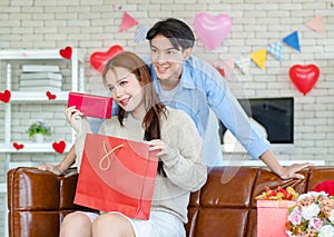 Asian young handsome male boyfriend standing smiling behind beautiful female girlfriend giving red wrapped present gift box in