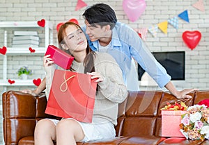 Asian young handsome male boyfriend standing smiling behind beautiful female girlfriend giving red wrapped present gift box in