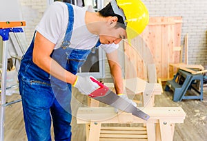 Asian young handyman and carpenter wearing yellow safety helmet and cutting wood by saw